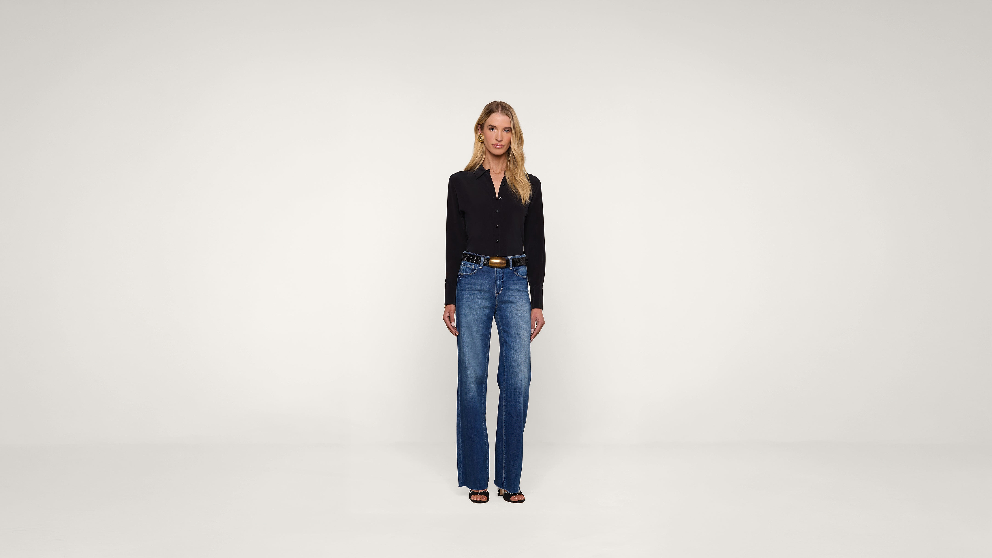 A woman stands facing forward in a plain, well-lit studio, wearing a black long-sleeve shirt, blue jeans, a brown belt, and black open-toe heels. The background is white and uncluttered.