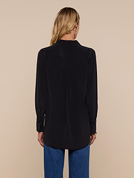 A woman with long blonde hair is standing with her back to the camera, wearing the Babette Silk Blouse in black and blue jeans against a plain, light background.