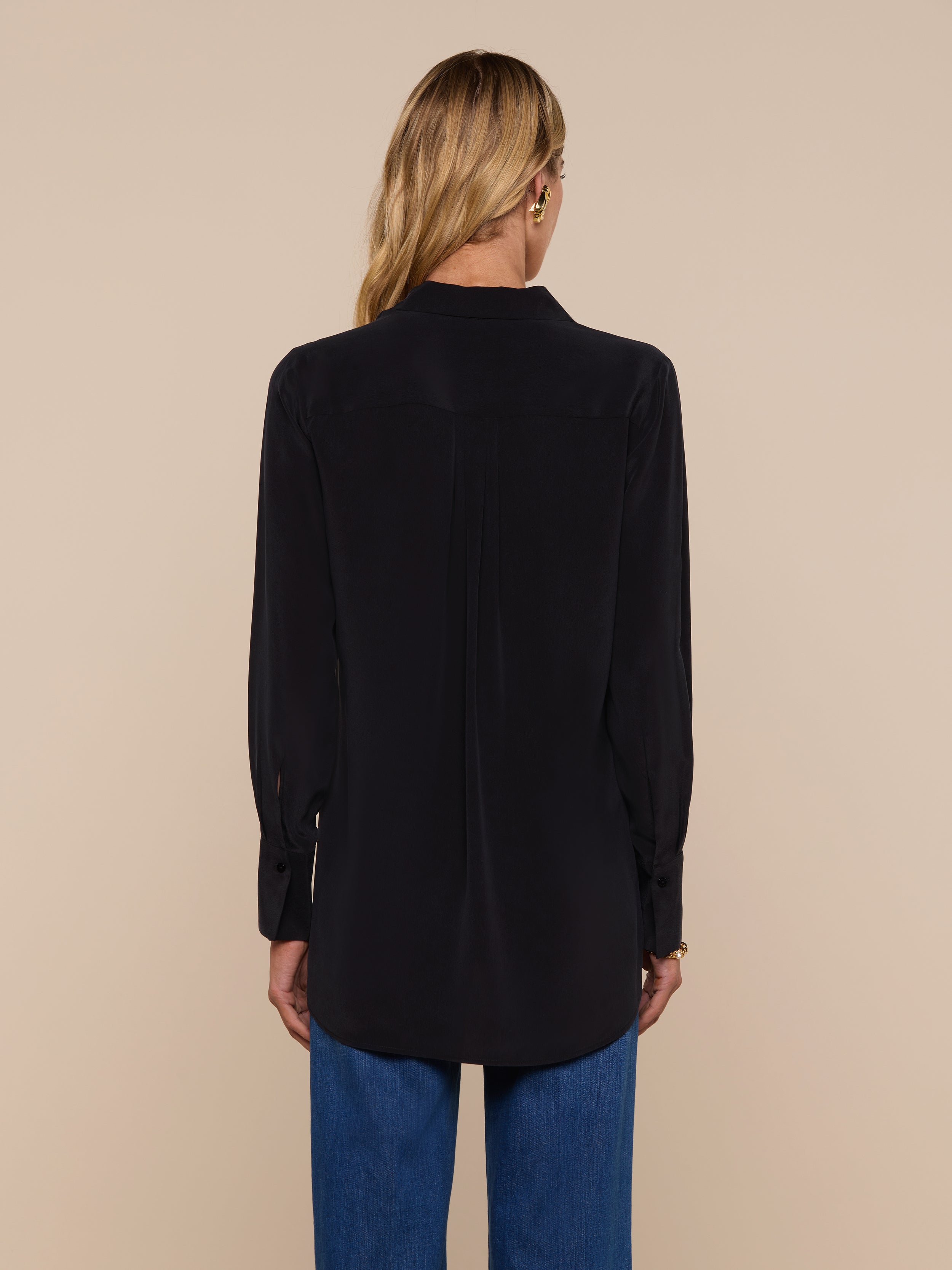 A woman with long blonde hair is standing with her back to the camera, wearing the Babette Silk Blouse in black and blue jeans against a plain, light background.