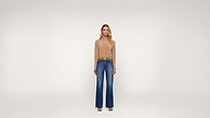 A woman stands facing forward in a beige long-sleeve top, blue wide-leg jeans, a brown belt, and sandals, against a plain white background.