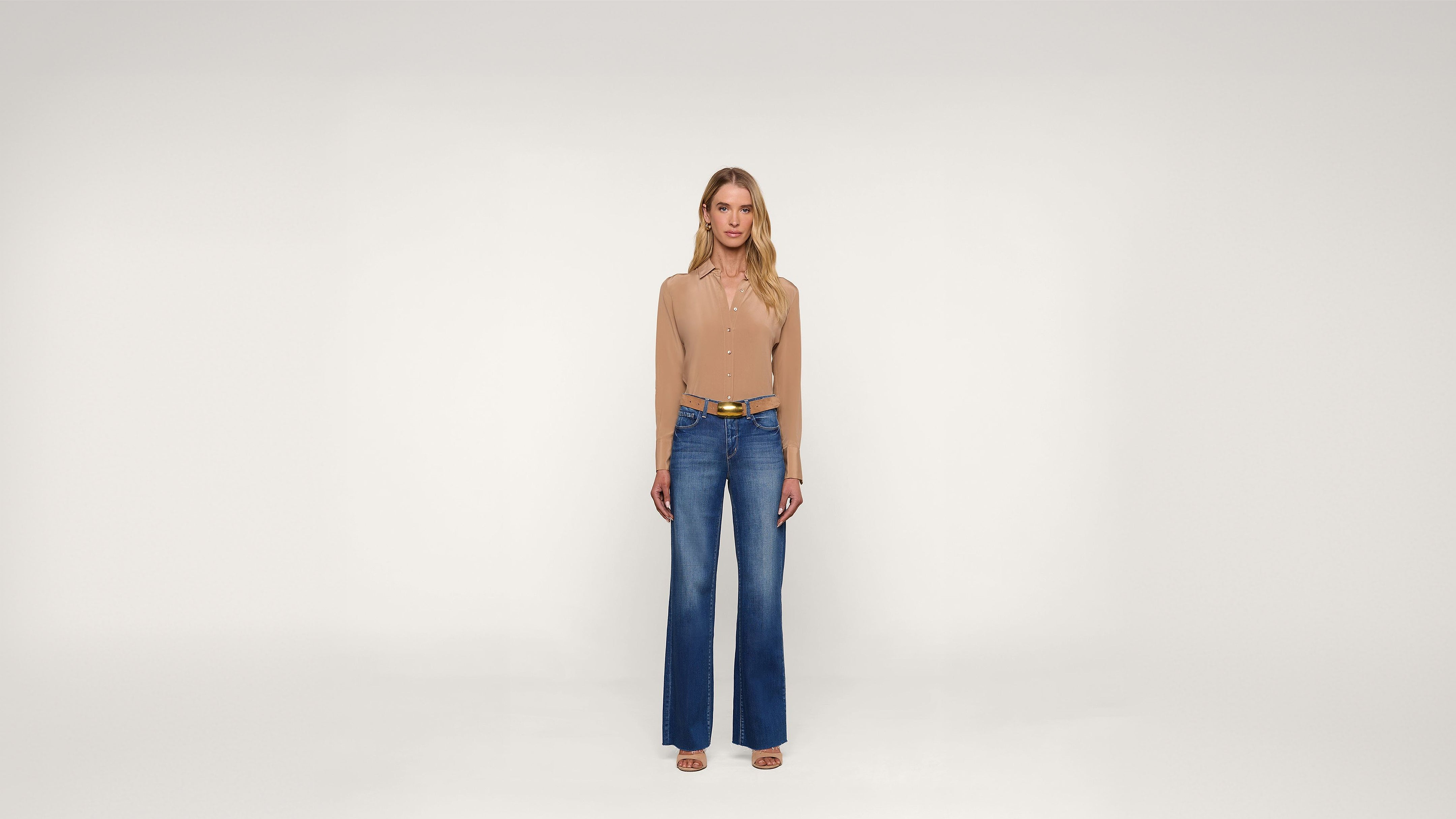A woman stands facing forward in a beige long-sleeve top, blue wide-leg jeans, a brown belt, and sandals, against a plain white background.