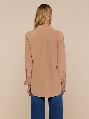 A woman with long blonde hair is seen from the back, wearing the Babette Silk Blouse in tan—a relaxed fit, long-sleeve top with a curved hem that falls below the hips—paired with blue jeans against a neutral beige background.