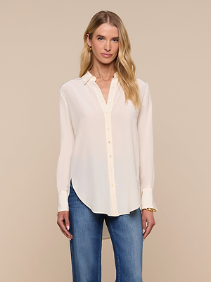 A woman with long blonde hair wears the Babette Silk Blouse, a relaxed-fit cream crepe de chine top, paired with blue jeans, standing against a beige background.