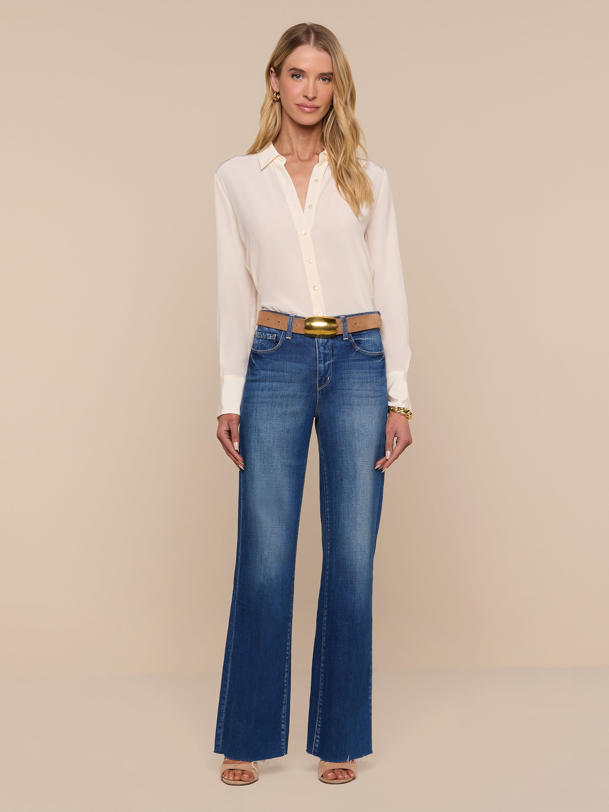 A woman stands against a plain white background wearing the Babette Silk Blouse in light pink, paired with blue flared jeans with a relaxed fit, a brown belt, and open-toed heels. She has long blonde hair and is smiling slightly.