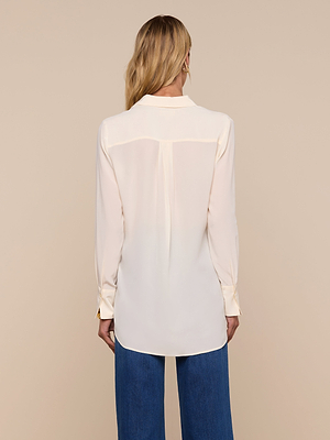 A person with long blonde hair, wearing the Babette Silk Blouse and blue pants, stands with their back to the camera against a beige background.