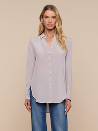 A woman wearing the Babette Silk Blouse, a light gray button-up with long sleeves and a crepe de chine finish, paired with blue jeans, stands facing forward against a plain, light background.