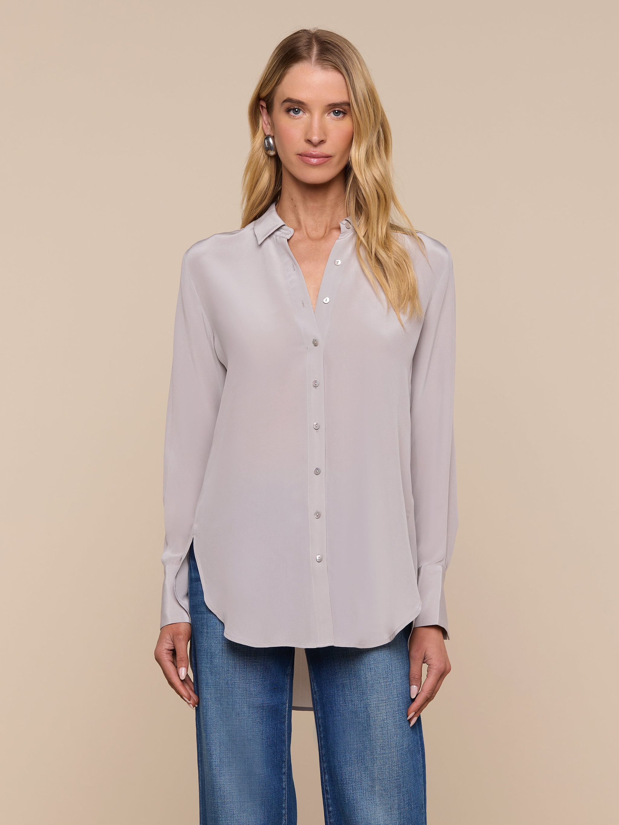 A woman wearing the Babette Silk Blouse, a light gray button-up with long sleeves and a crepe de chine finish, paired with blue jeans, stands facing forward against a plain, light background.