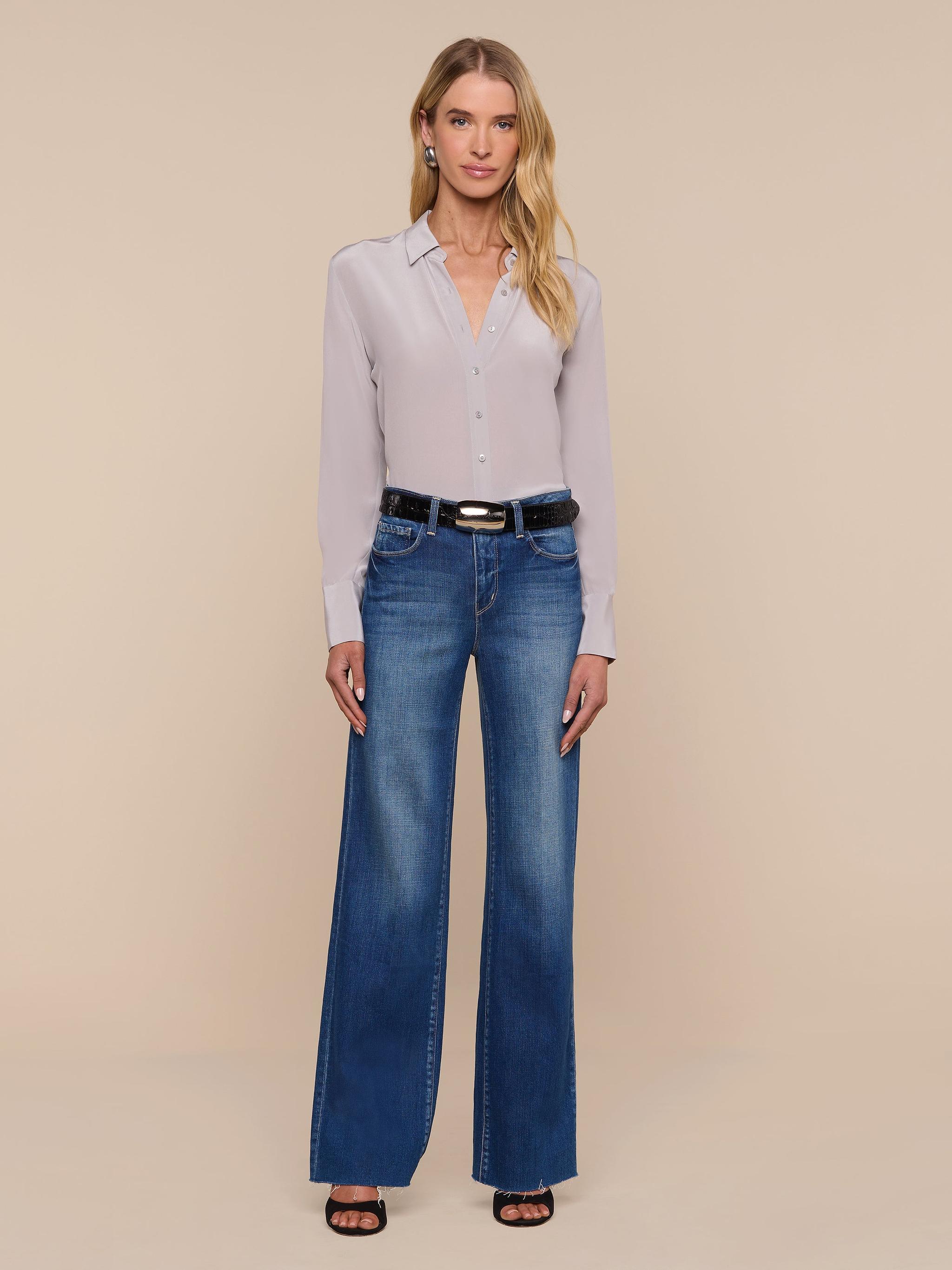 A woman stands against a white background, wearing the Babette Silk Blouse—a light gray crepe de chine button-up with a relaxed fit—tucked into blue wide-leg jeans, styled with a black belt and black heeled sandals. She has long blonde hair and is facing the camera.