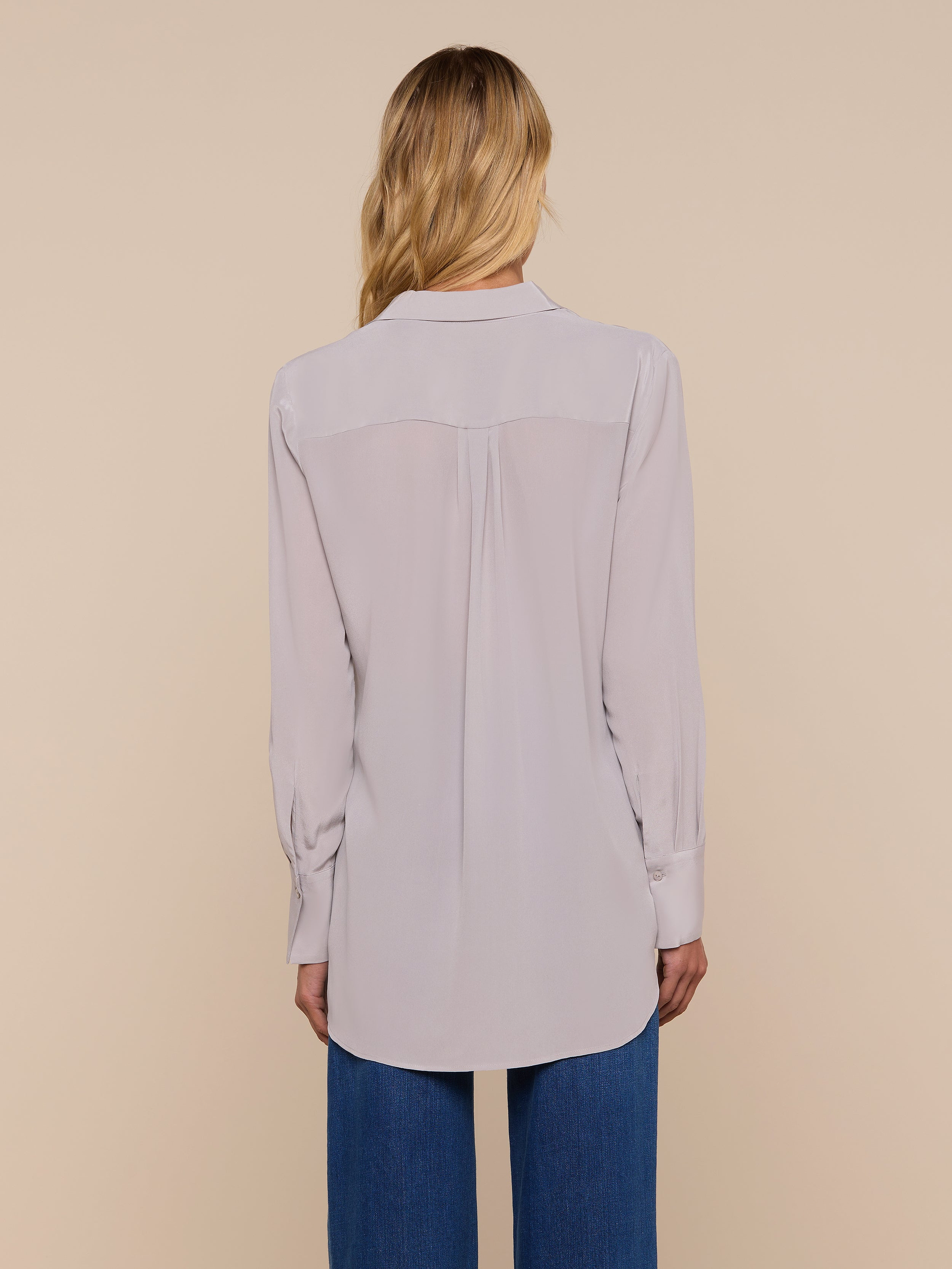 A woman with blonde hair is shown from behind, wearing the Babette Silk Blouse in light gray with a relaxed fit and blue jeans, standing against a plain off-white background.