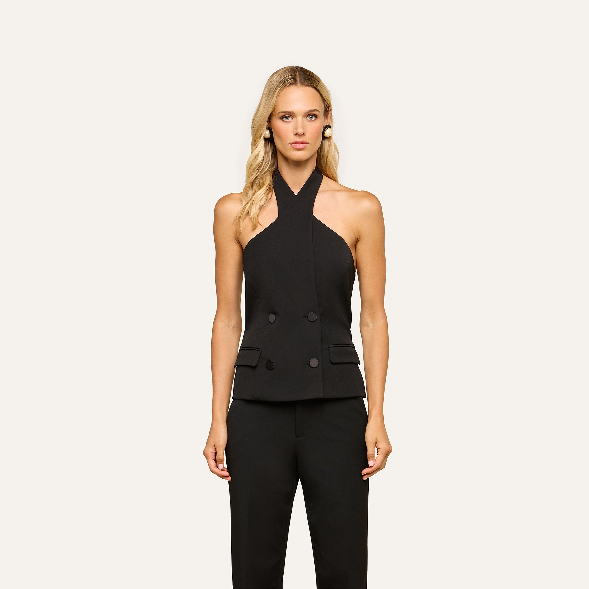 A woman stands against a plain light background, wearing the sleeveless black Rhoda Top with double-breasted buttons and matching black pants.