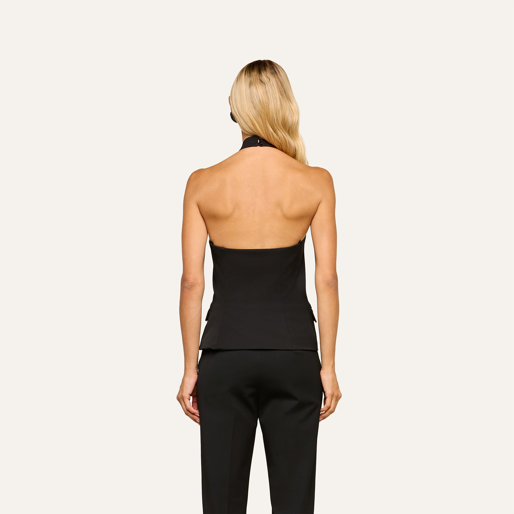 A woman with long blonde hair is shown from the back, wearing the Rhoda Top and black pants, standing against a plain light background.