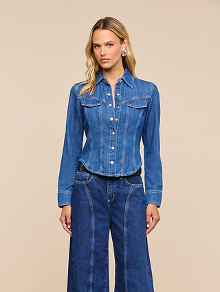 The woman wears the Louis Fitted Shirt, a blue denim jacket with buttons, paired with matching wide-leg jeans as she stands against a plain beige background. Her long blonde hair is styled simply, and she gazes straight ahead with a neutral expression, embodying equestrian sophistication.