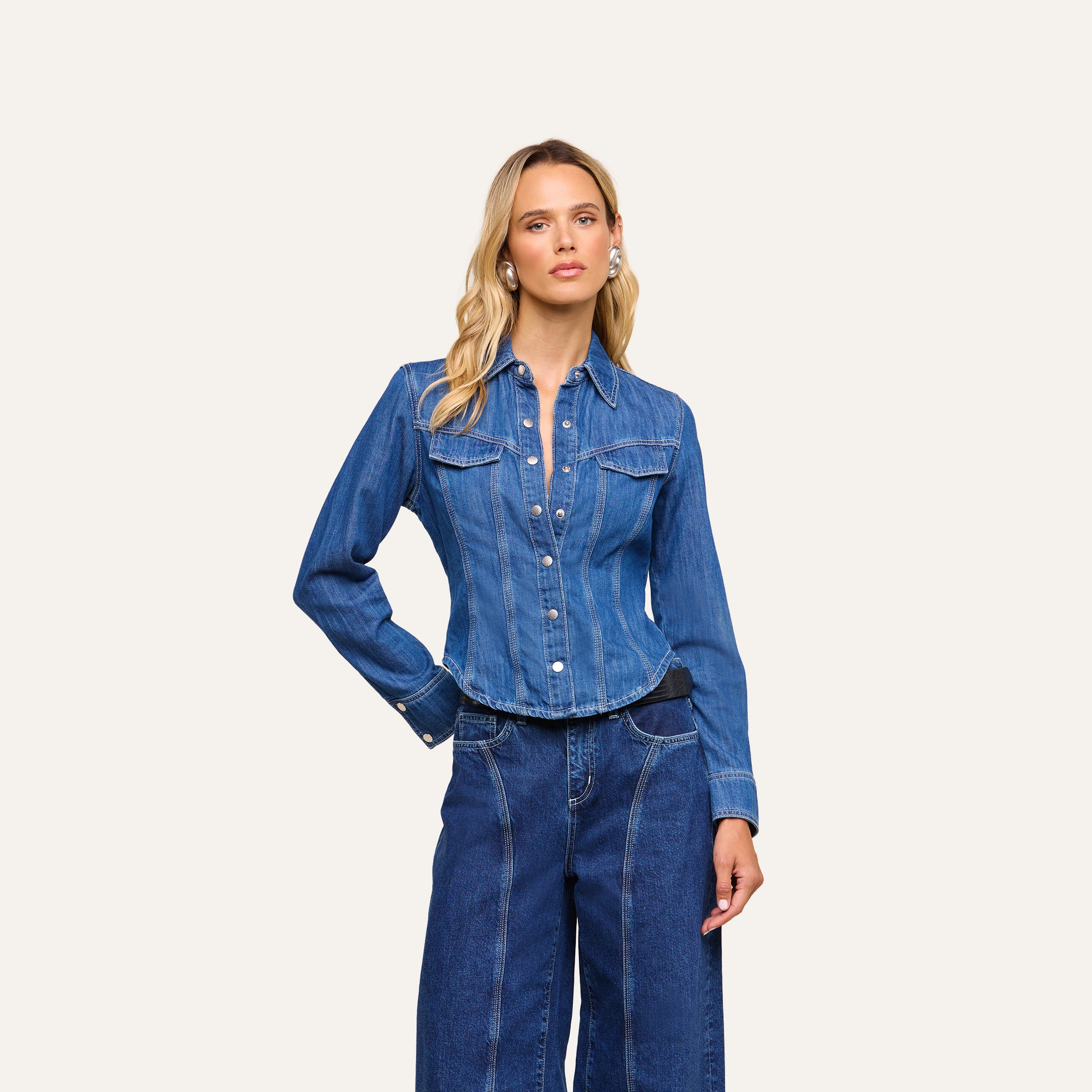 Dressed in the Louis Fitted Shirt with long sleeves and paired with wide-leg high-waisted jeans, she stands against a plain light background, radiating equestrian sophistication.