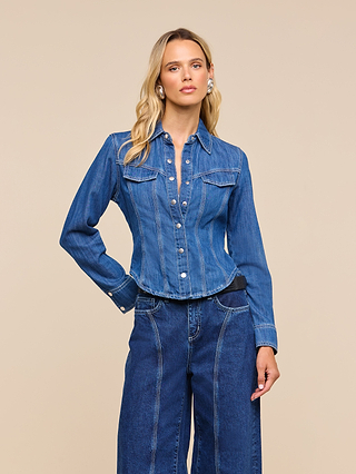 A woman wearing the Louis Fitted Shirt paired with wide-leg denim jeans stands against a plain beige background, gazing confidently at the camera—her poised stance capturing a sense of equestrian sophistication.