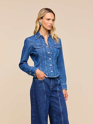 A woman with long blonde hair wears the Louis Fitted Shirt, an ultra light denim button-up, paired with wide-leg jeans as she stands against a plain beige background.