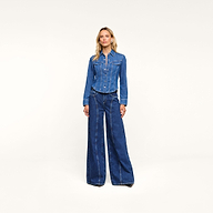 A woman stands against a plain white background, wearing the Louis Fitted Shirt and wide-leg blue denim jeans, with her hands by her sides, capturing an air of equestrian sophistication.