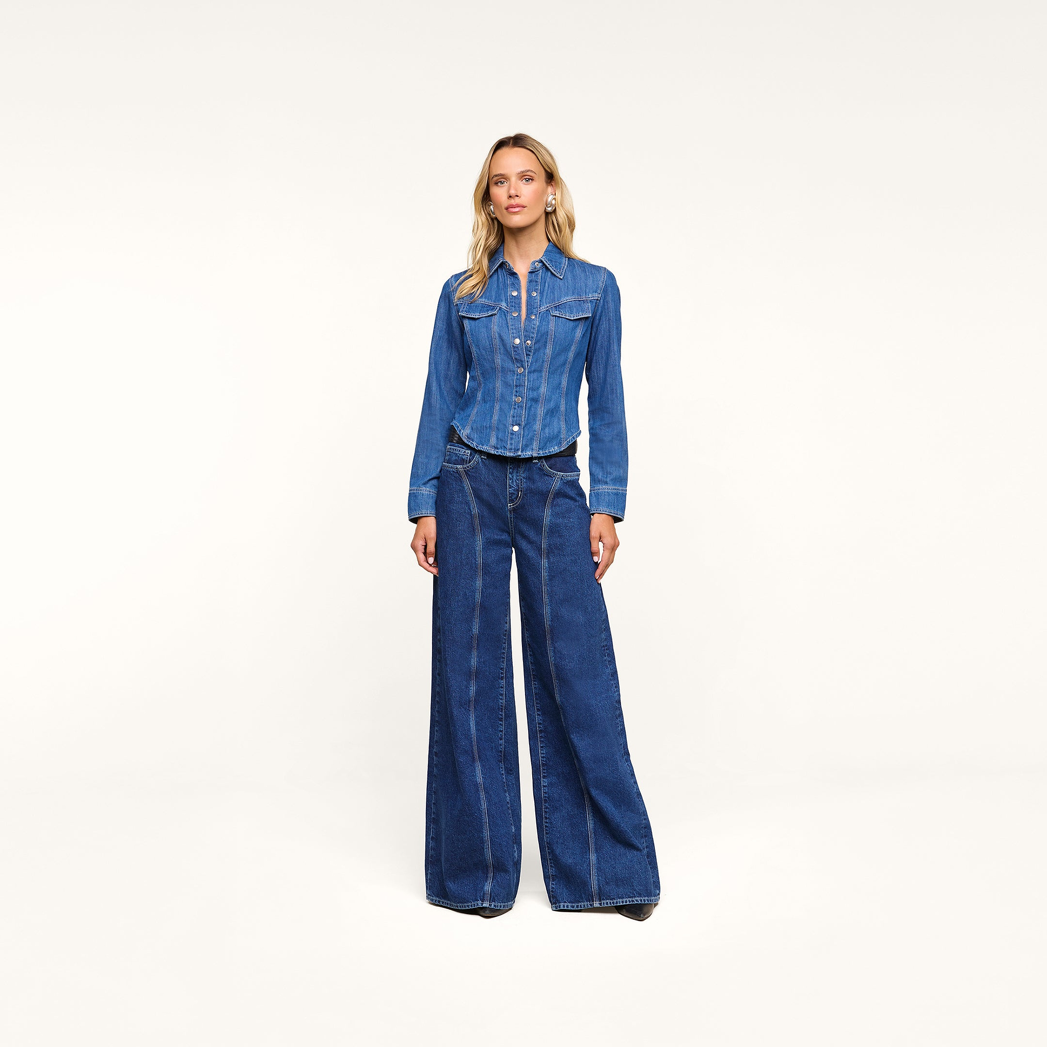 A woman stands against a plain white background, wearing the Louis Fitted Shirt and wide-leg blue denim jeans, with her hands by her sides, capturing an air of equestrian sophistication.