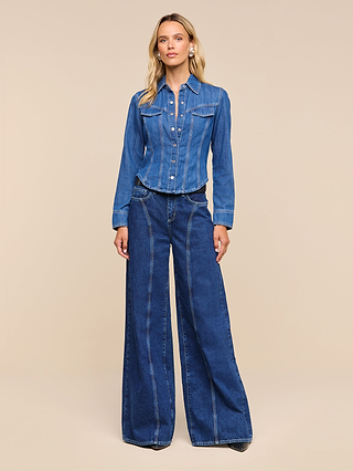 Woman standing against a beige background, embodying equestrian sophistication in the Louis Fitted Shirt and wide-leg blue denim pants, with hands relaxed by her sides.