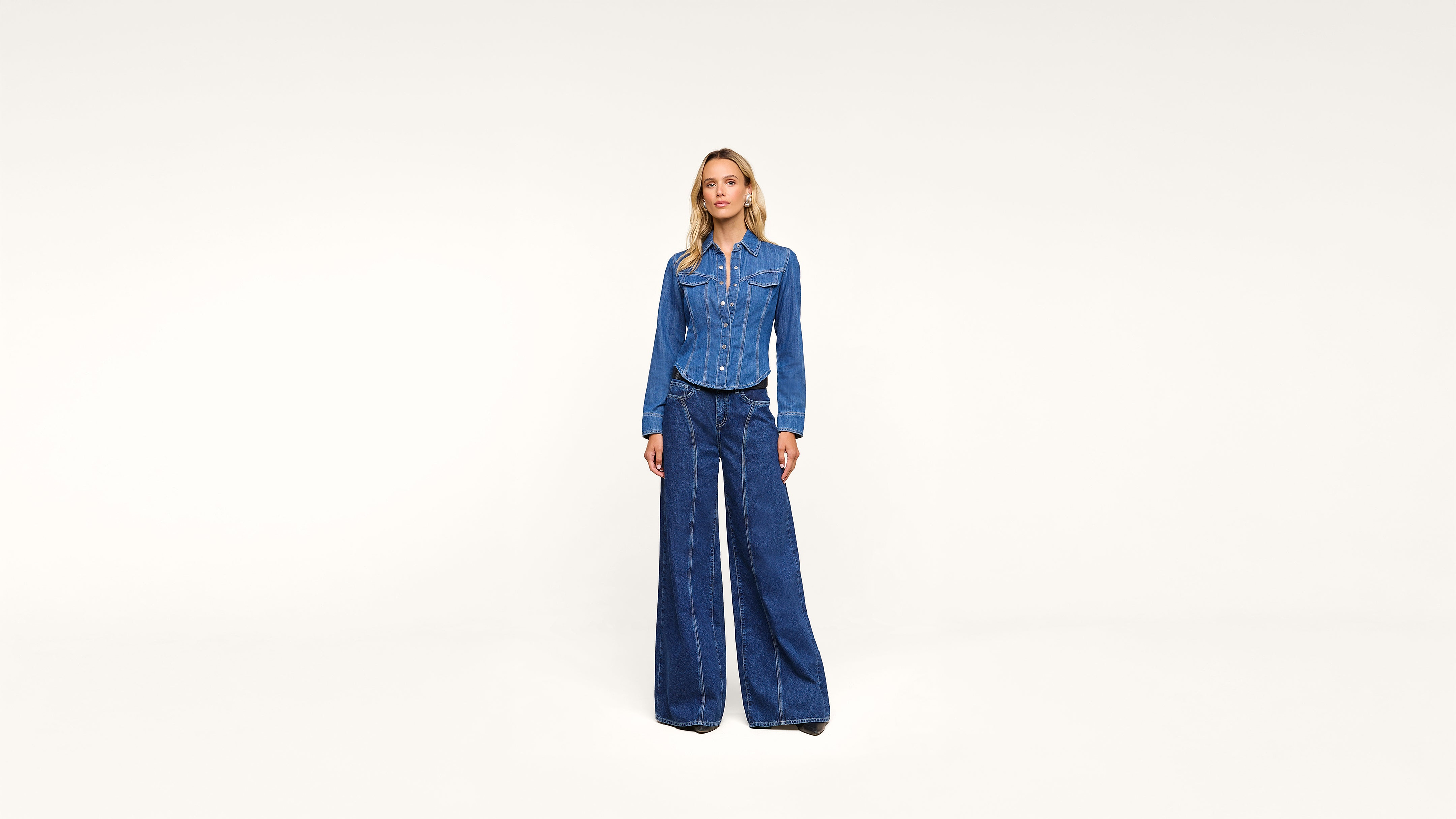 A woman stands against a plain white background, wearing a blue denim button-up jacket and matching wide-leg denim pants.