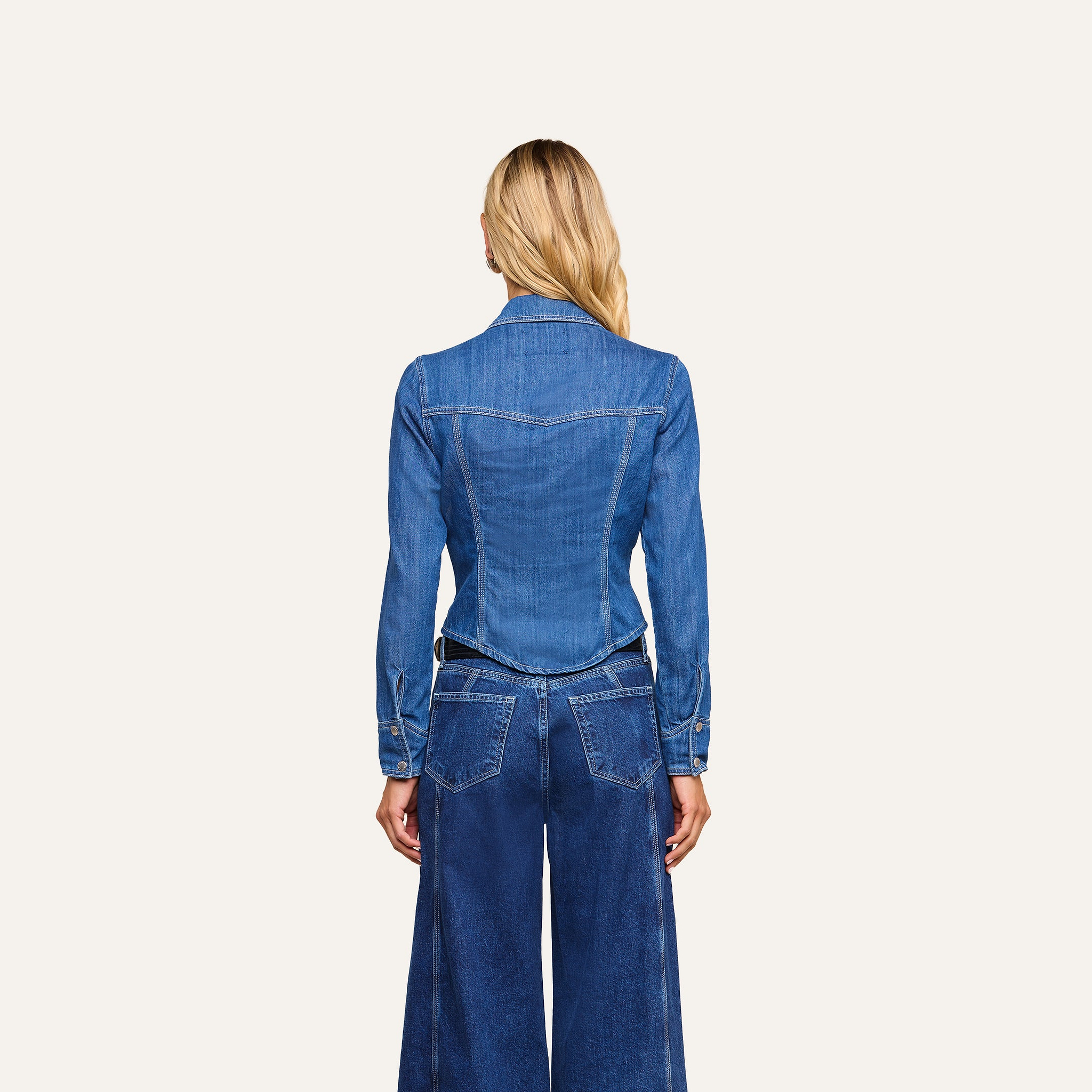 A person with long blonde hair is shown from the back, capturing equestrian sophistication in the Louis Fitted Shirt and wide-leg blue jeans, standing against a plain light background.