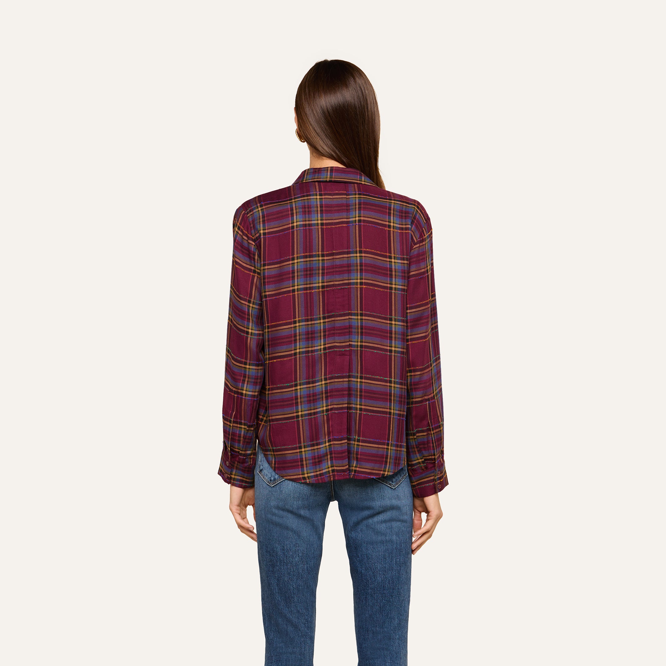 A woman stands with her back to the camera against a plain light background, wearing the Suzette Shirt—a red plaid button-down with a high-low hemline—paired with blue jeans.