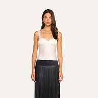A woman with long, wavy brown hair stands against a plain background, wearing the Soleia Lace Camisole in white silk charmeuse with delicate lace straps, paired with a high-waisted black skirt adorned with long fringe detailing. She is facing forward.