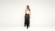 A woman wearing sunglasses, a white fitted top, and a black fringed skirt with black boots stands against a plain white background.