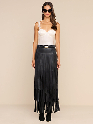 Wearing sunglasses and the Soleia Lace Camisole, featuring eyelash lace straps, paired with a long black fringed skirt and belt, a woman stands against a plain beige background.