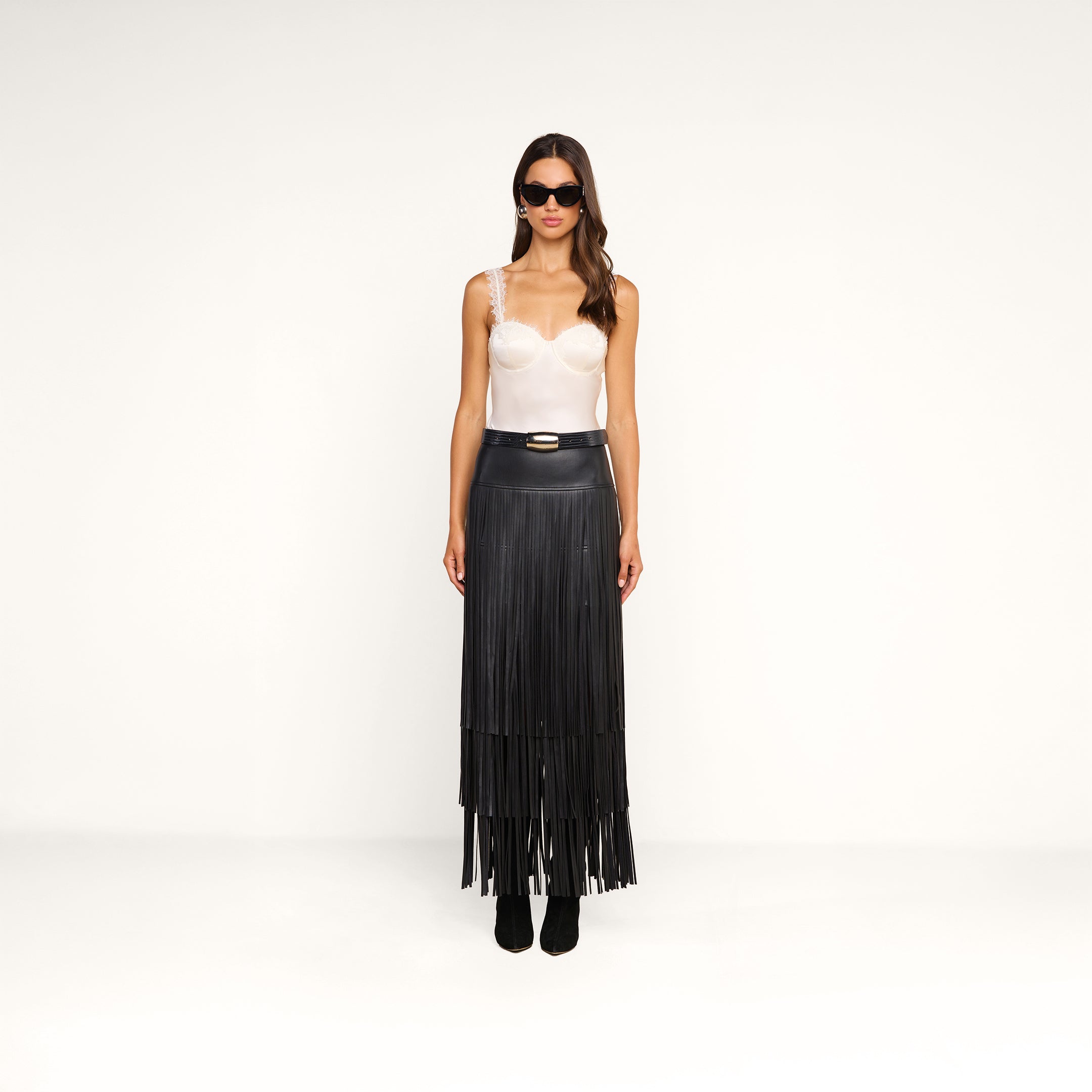 Woman wearing sunglasses, a white Soleia Lace Camisole, and a high-waisted long black fringe skirt with black ankle boots, standing against a plain white background.
