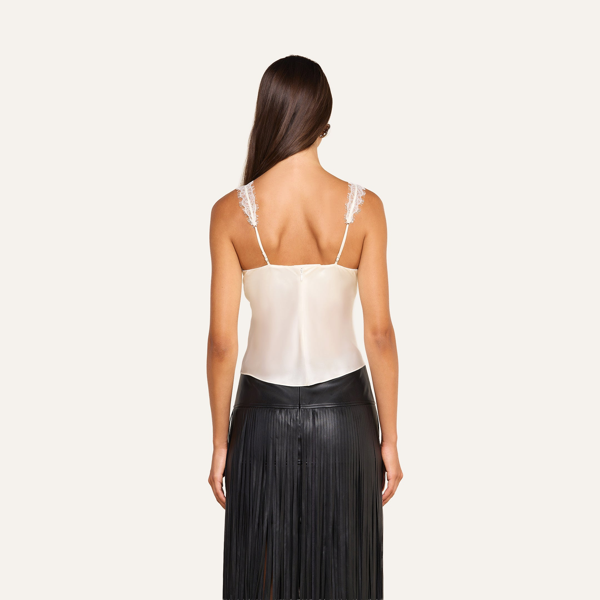 A woman is shown from the back wearing a white Soleia Lace Camisole with lace straps and a black pleated leather skirt. Her brown hair is long and straight, and she stands against a plain, light background.