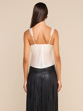 A woman with long brown hair is shown from the back against a beige background, wearing the Soleia Lace Camisole—a white silk top with delicate eyelash lace straps—paired with a black pleated skirt.