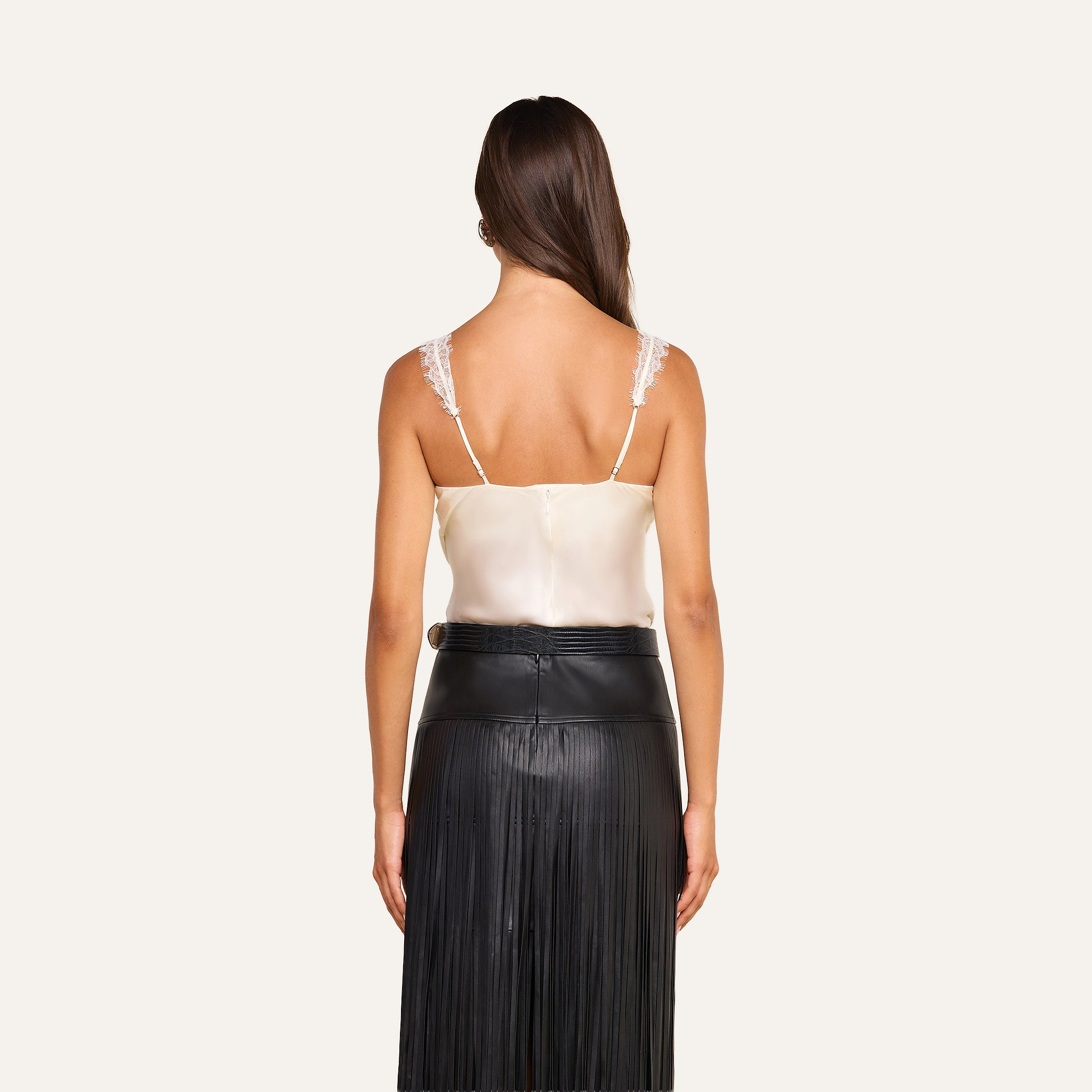A woman stands facing away, wearing the cream Soleia Lace Camisole—crafted from silk charmeuse with lace straps—and a black high-waisted skirt featuring a fringed design. The background is plain and light-colored.