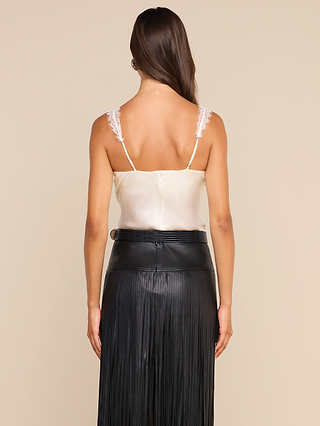 A woman stands facing away, wearing the Soleia Lace Camisole in cream with delicate eyelash lace trim, paired with a high-waisted black leather skirt featuring a long, fringed design. Her brown hair is down against a beige background.