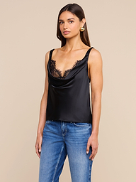 A woman wearing the Evadne Silk Lace Camisole in black with lace trim, paired with blue jeans, stands against a plain light background, looking slightly to the side with a neutral expression.