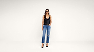 Woman in sunglasses wearing a black camisole, blue jeans with a knee rip, and black heels stands against a plain white background.