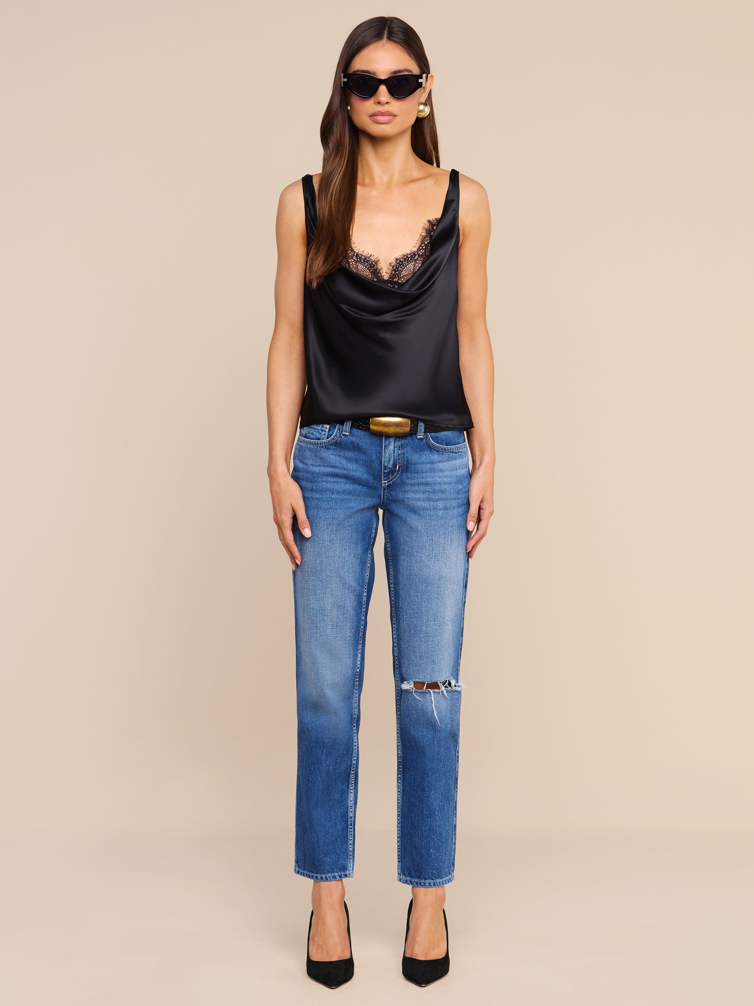 A woman wearing sunglasses, the Evadne Silk Lace Camisole, blue jeans with a rip on one knee, and black heels stands facing forward against a plain white background.