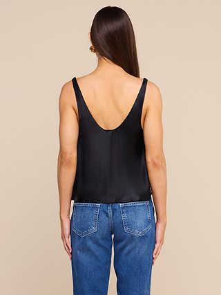 A person with long brown hair, wearing a sleeveless black top reminiscent of the Evadne Silk Lace Camisole and blue jeans, is standing facing away from the camera against a beige background.