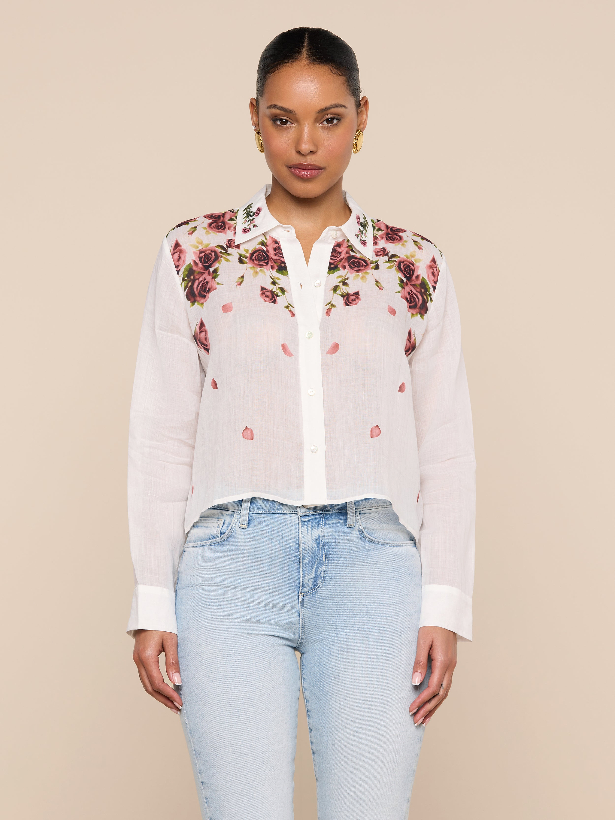 A woman stands facing forward, wearing the Philomena Blouse—a semi-sheer ramie button-down shirt adorned with colorful floral embroidery on the shoulders—paired with light blue jeans, against a plain light background.