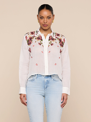 A woman wears the Philomena Blouse—a white Ramie button-down with rose print embroidery on the collar and shoulders—paired with light blue jeans, standing facing forward against a plain beige background.