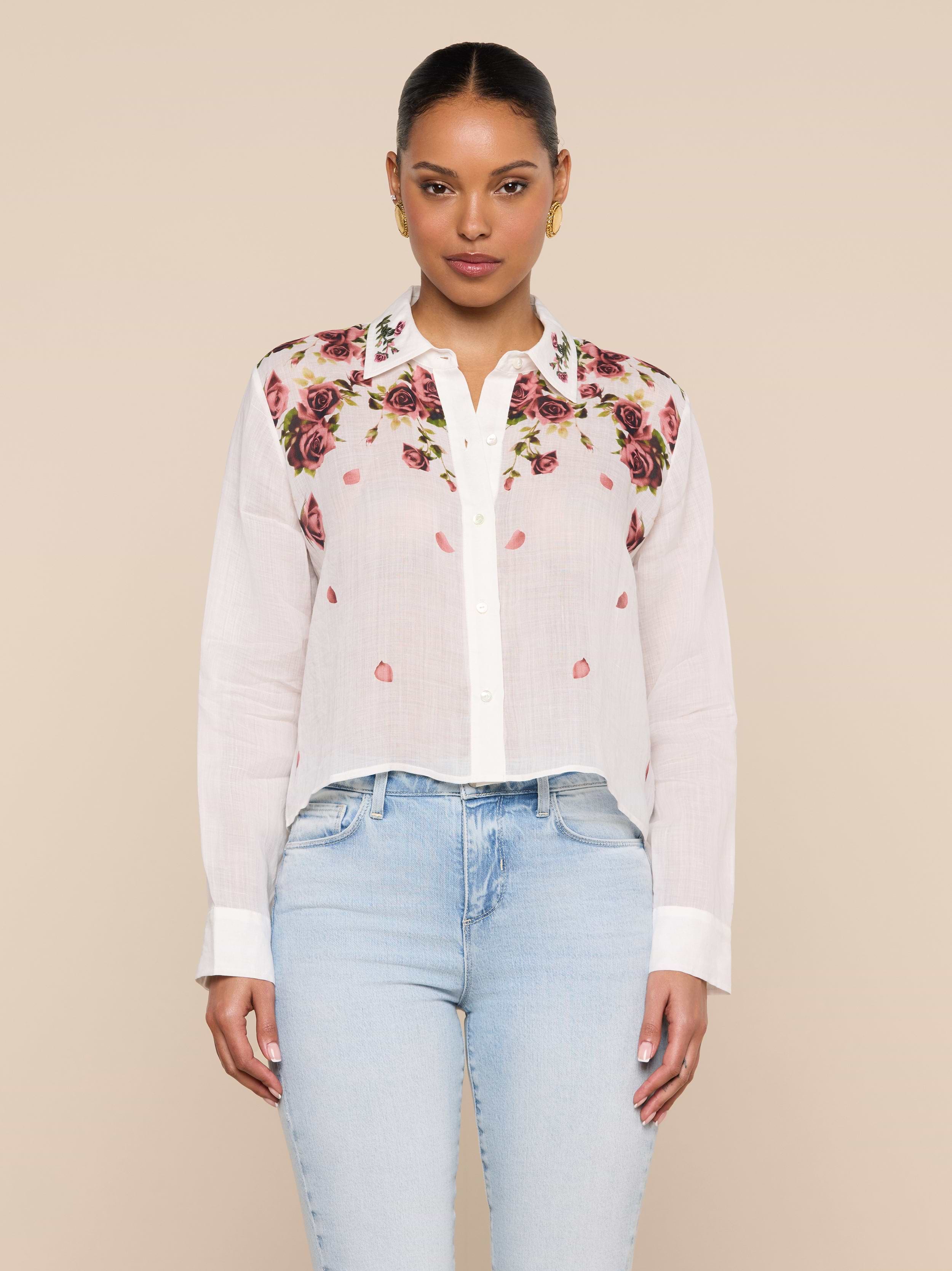 A woman stands facing forward, wearing the Philomena Blouse—a semi-sheer ramie button-down shirt adorned with colorful floral embroidery on the shoulders—paired with light blue jeans, against a plain light background.