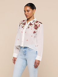 A woman wearing the Philomena Blouse—a semi-sheer ramie button-down shirt with floral embroidery on the shoulders and collar—paired with light blue jeans, stands against a plain light background.