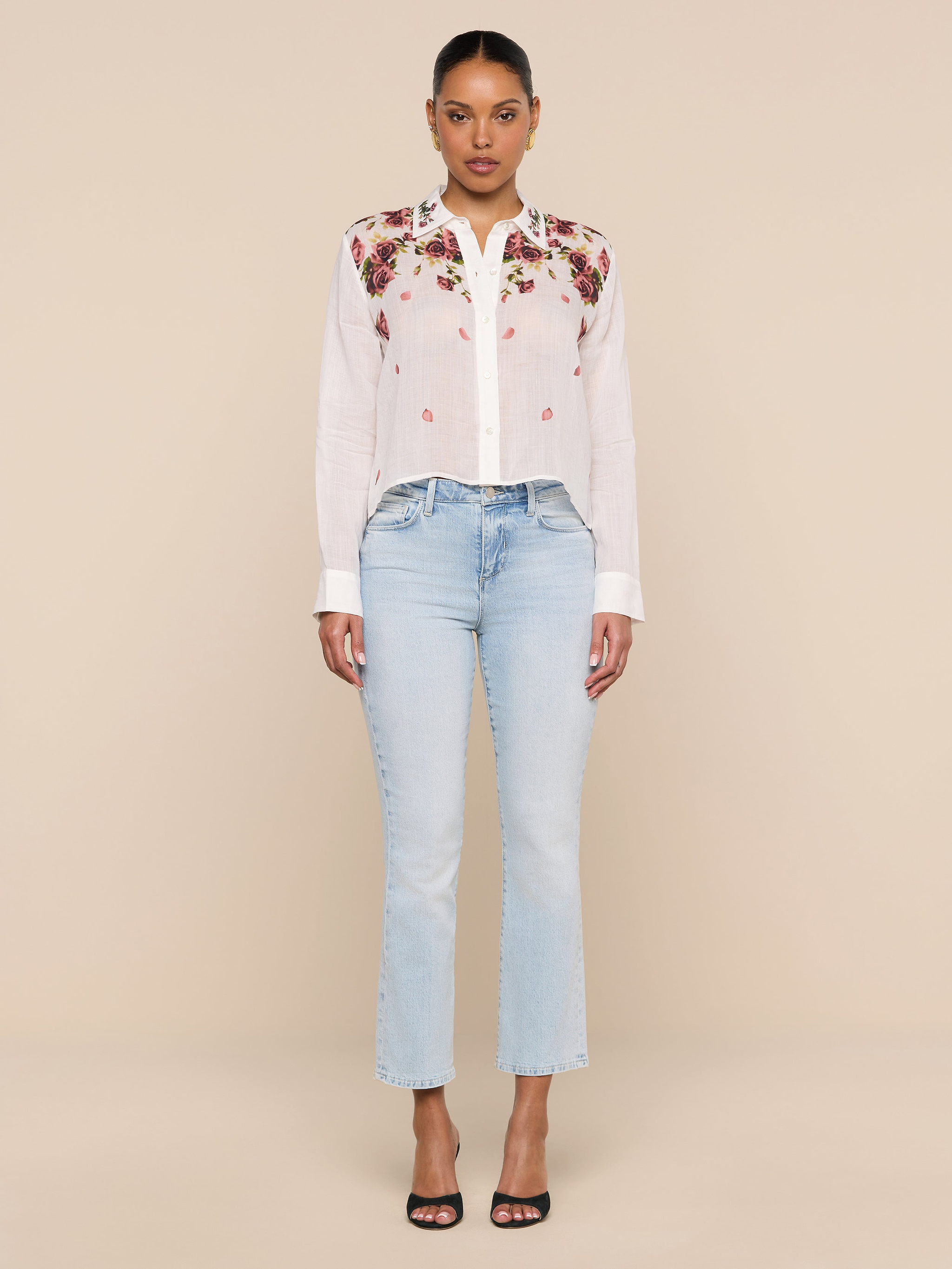 A woman stands facing forward against a plain white background wearing the Philomena Blouse, a semi-sheer white Ramie top with floral embroidery, paired with light blue jeans and black heeled sandals.