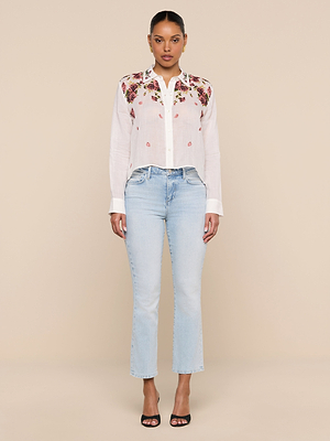 A woman stands against a beige background, wearing the Philomena Blouse—white ramie with floral shoulder embroidery—paired with light blue jeans and black open-toe heels. She faces forward, arms relaxed at her sides.