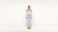A woman stands facing forward against a plain white background, wearing a white blouse with floral embroidery, light blue jeans, and black heeled sandals.