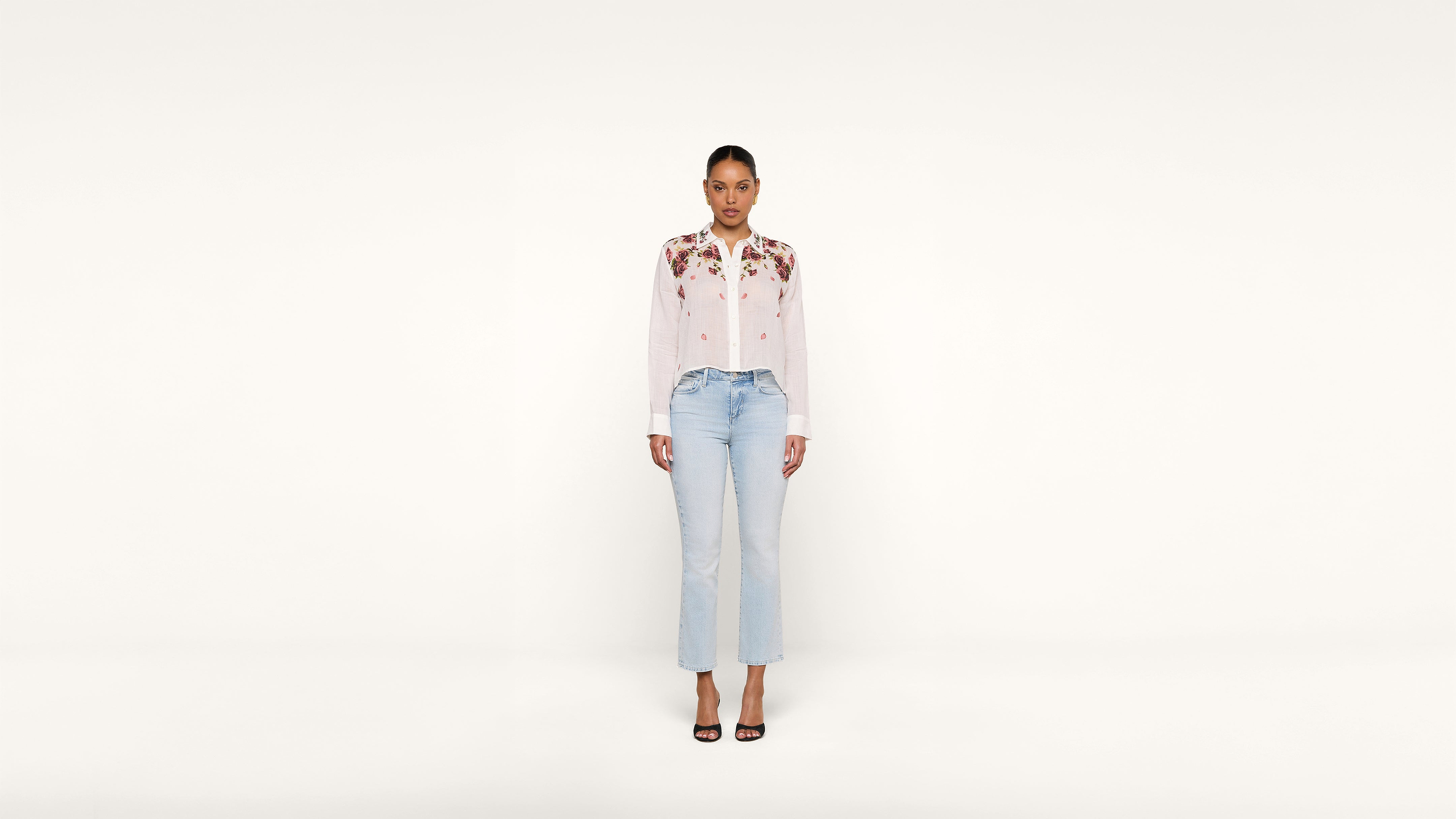 A woman stands facing forward against a plain white background, wearing a white blouse with floral embroidery, light blue jeans, and black heeled sandals.