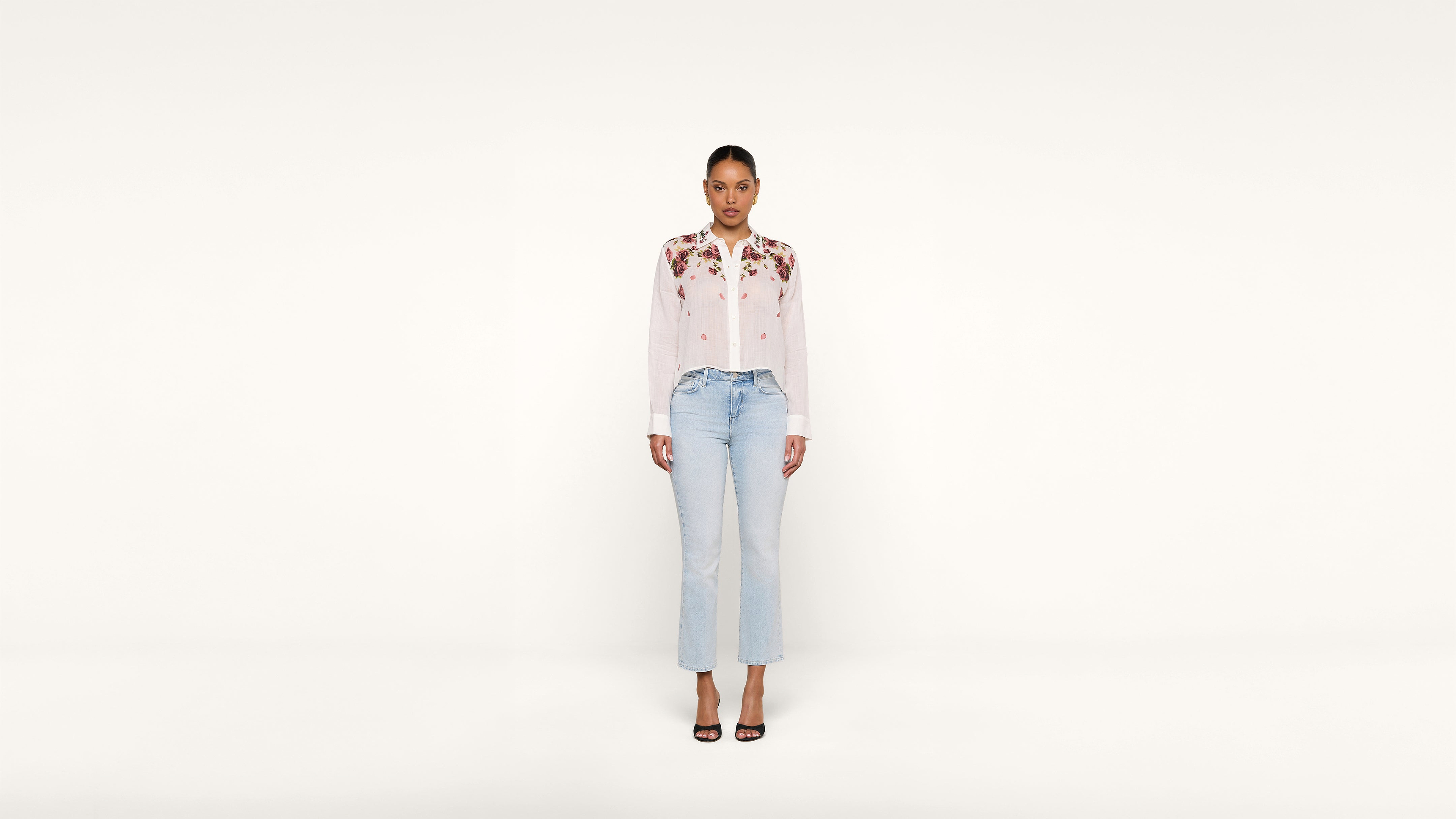 A woman stands facing forward against a plain white background, wearing a white blouse with floral embroidery, light blue jeans, and black heeled sandals.