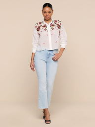 A woman stands against a white background wearing the semi-sheer Philomena Blouse with floral embroidery, light blue jeans, and black heels.