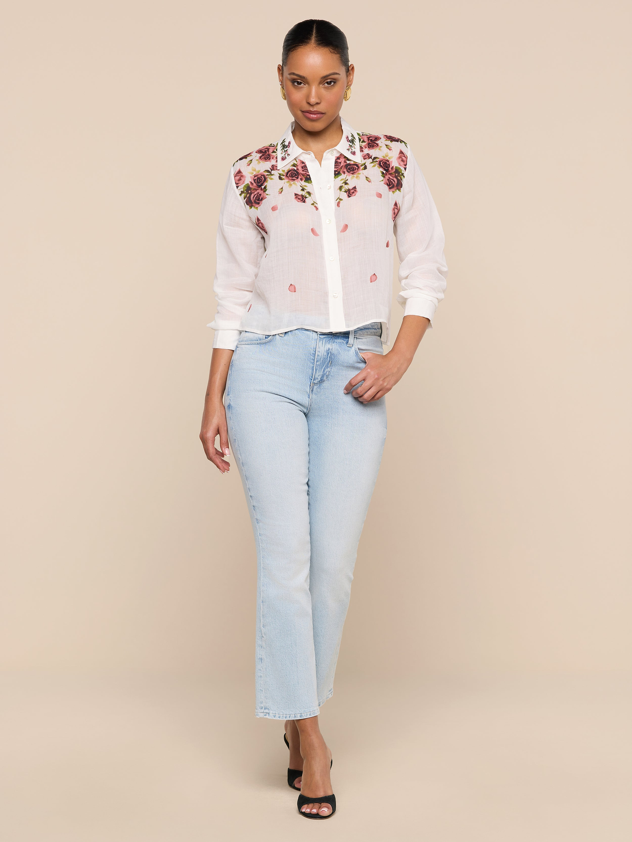 A woman stands against a white background wearing the semi-sheer Philomena Blouse with floral embroidery, light blue jeans, and black heels.