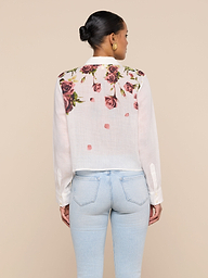 A woman stands facing away, wearing the Philomena Blouse—a white button-down shirt featuring a rose print on the upper back—paired with light blue jeans. Her hair is styled in a bun and she accessorizes with gold hoop earrings.