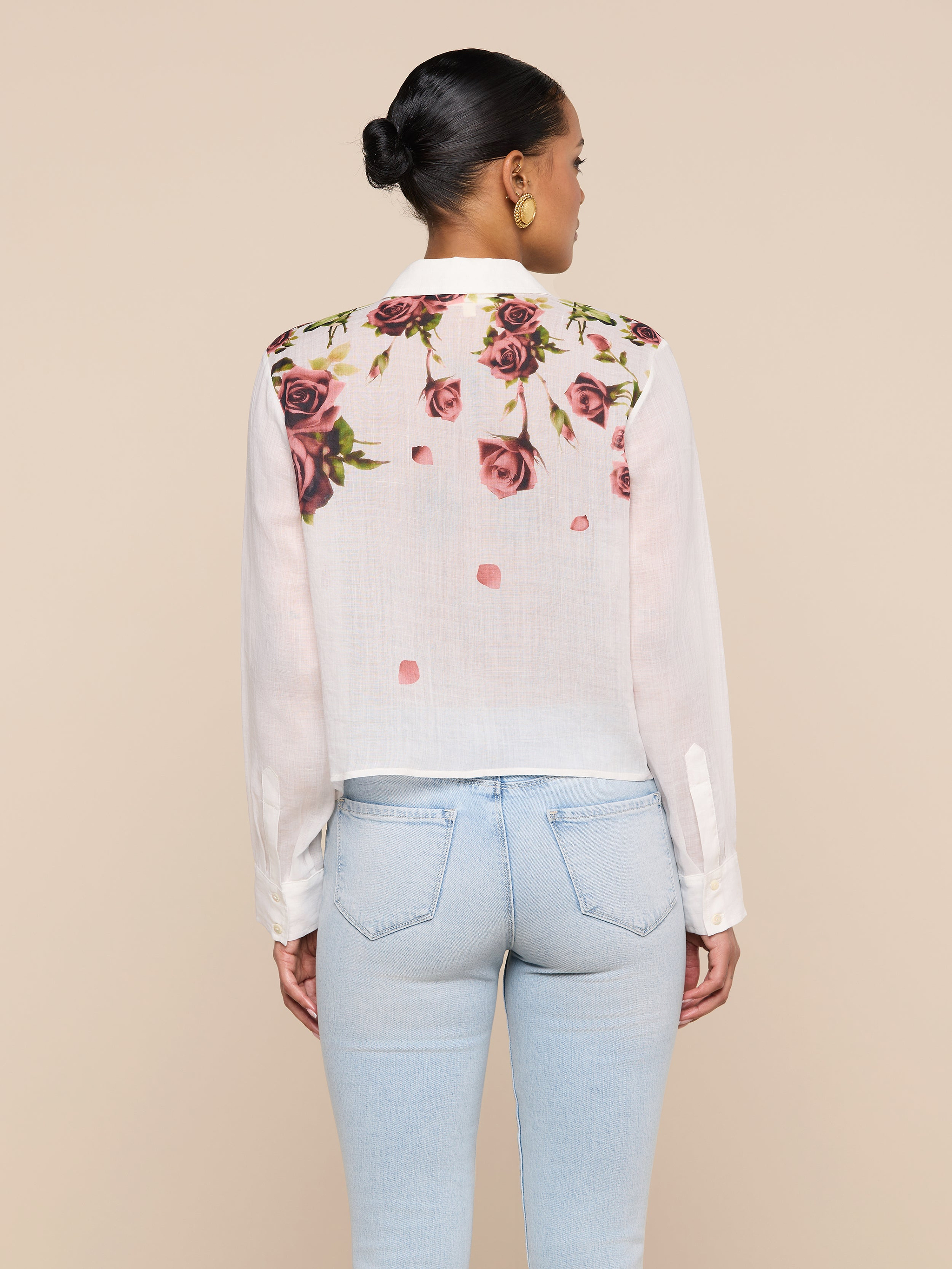 A woman stands facing away, wearing the Philomena Blouse—a white button-down shirt featuring a rose print on the upper back—paired with light blue jeans. Her hair is styled in a bun and she accessorizes with gold hoop earrings.